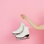 A close-up of a hand holding white ice skating shoes against a pink backdrop.
