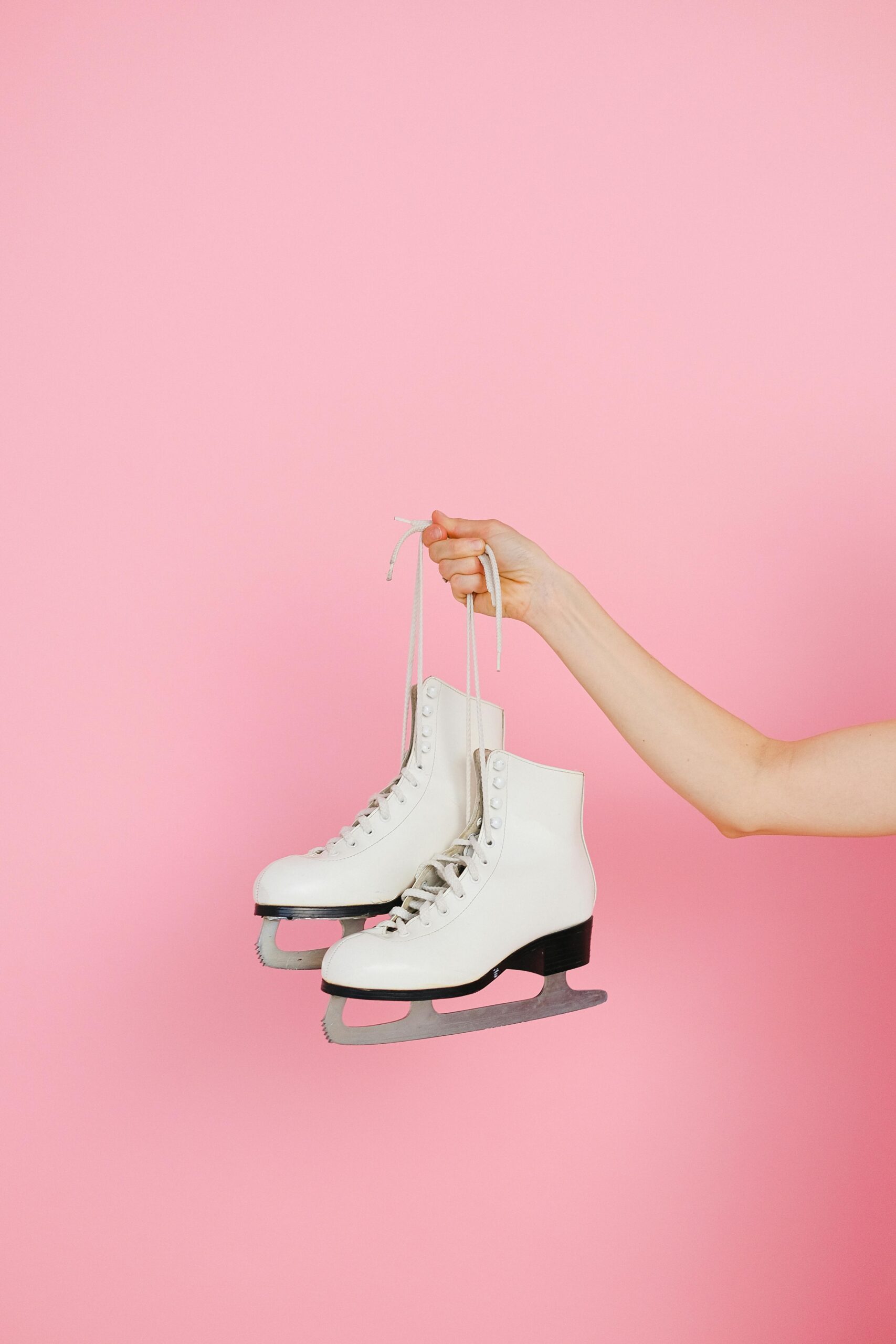 A close-up of a hand holding white ice skating shoes against a pink backdrop.