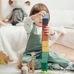 A cute child stacks colorful wooden blocks while playing indoors, surrounded by toys.